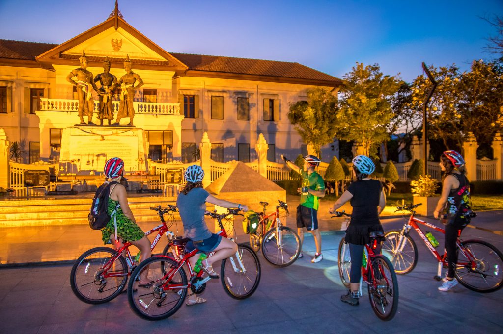 Guide and four cyclists stopped in front of Three Kings Monument in Chiang Mai, Thailand in evening light