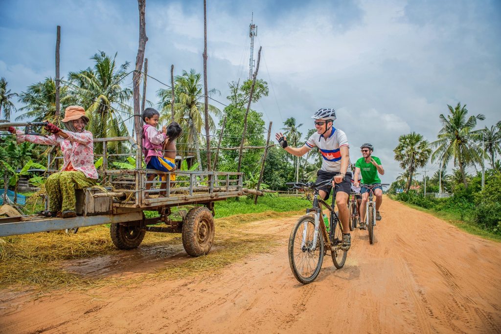 Two cyclists waving at local woman and boys on countryside road in Siem Reap
