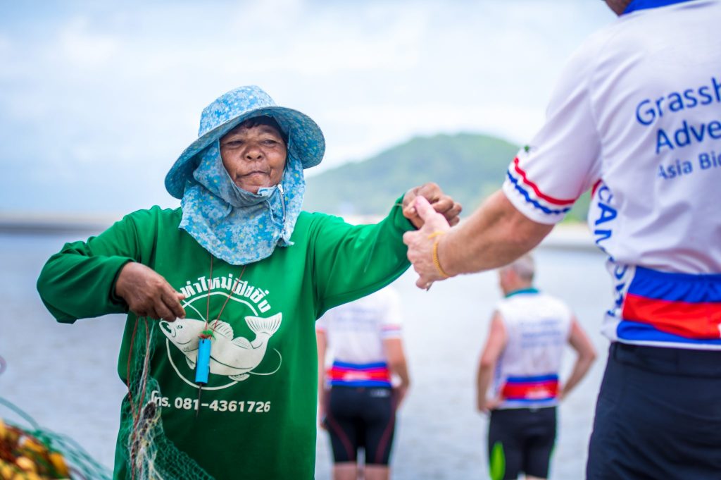 Local woman handing something to person in Grasshopper Adventures Thailand cycling jersey with water, two cyclists and hill behind