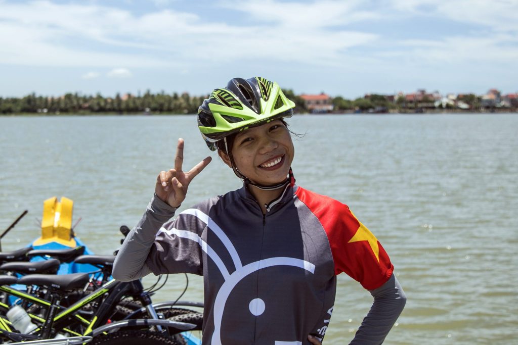 Smiling local leader in helmet and Grasshopper Adventures Vietnam cycling jersey with peace sign in front of water