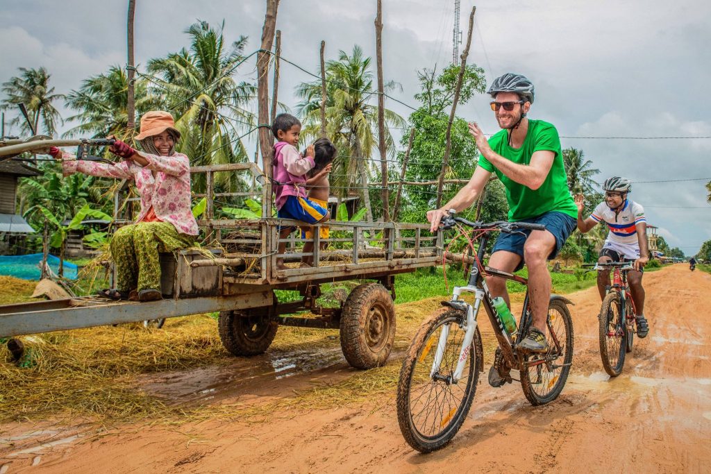 Two cyclists waving to local woman and children on red dirt road in Siem Reap